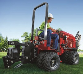 Worker in a white hard hat and safety vest operating a red ride-on trencher/vibratory plow machine on grass, with trees in the background